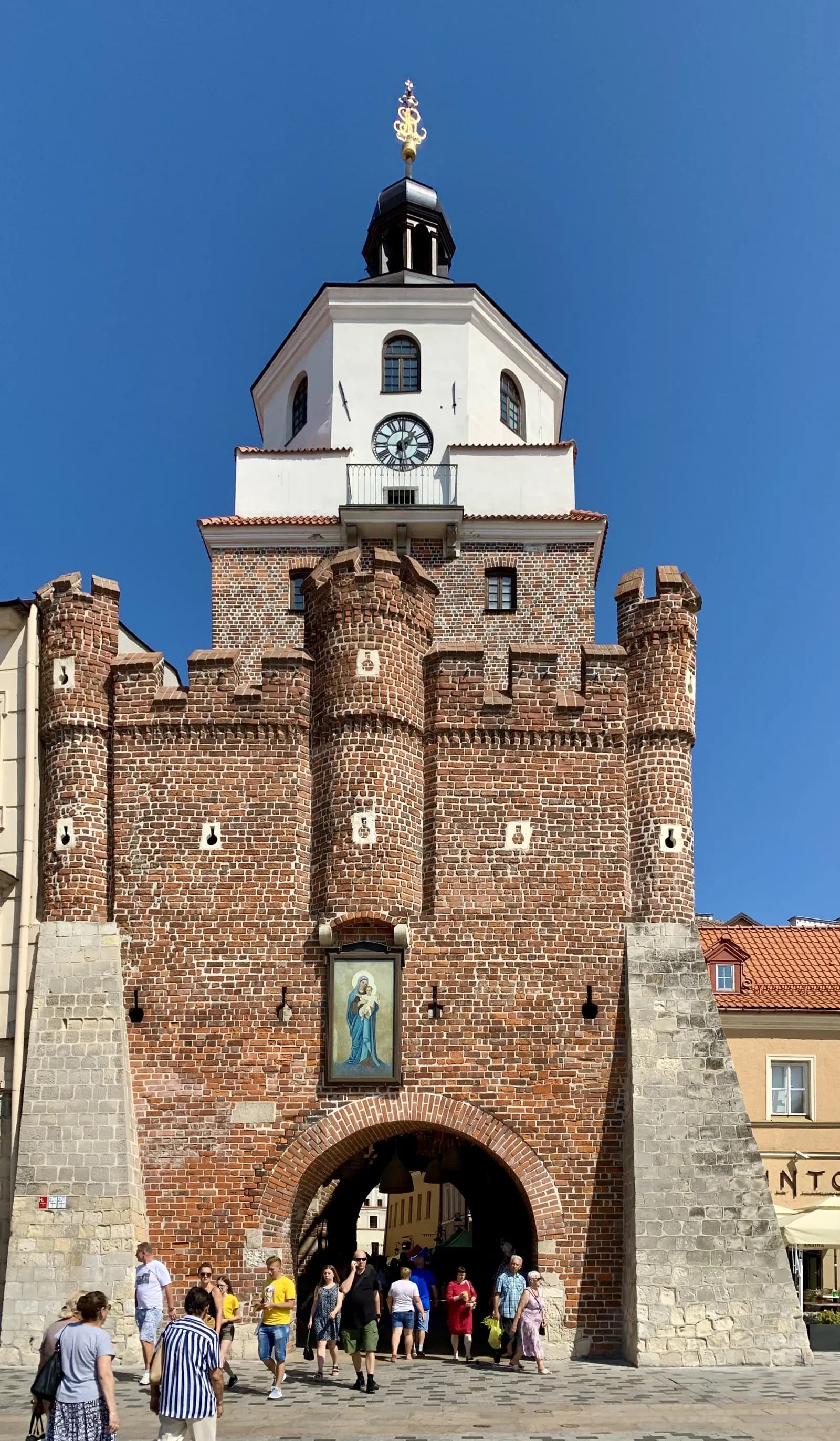 A tall brick gate with an archway at its base, topped with a white clock tower and golden spire, under a clear blue sky.